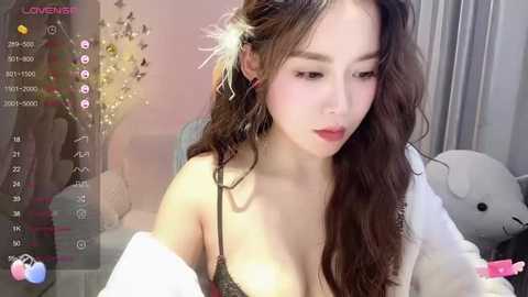 Media: Video of an Asian woman with long, wavy hair and fair skin, wearing a black lace bra and white robe. Soft lighting in a cozy room with a teddy bear and flowers in the background.