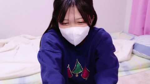 Media: Video of an East Asian woman in a blue sweatshirt with Christmas trees, wearing a white mask, lying on a bed with white sheets and a pink curtain in the background.