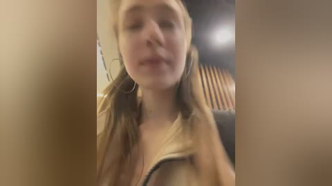 Media: A blurry video of a young woman with long, light brown hair and fair skin, wearing a beige jacket, looking slightly out of focus.