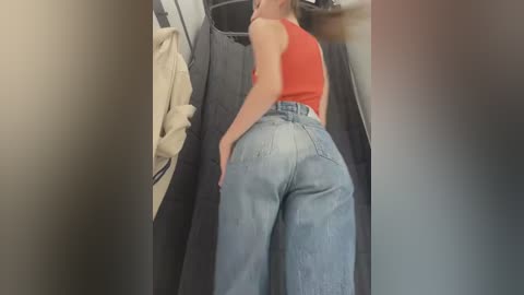 Media: Video of a woman with light skin, wearing a red tank top and high-waisted light blue jeans, standing in a fitting room. A beige coat hangs on the left.