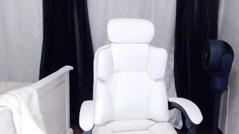 Media: Video of a modern, white leather gaming chair with high backrest and armrests, positioned in a minimalist room with black curtains and a white wall. A blue computer speaker is visible on the right.