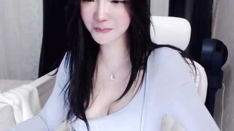 Media: Video of a pale-skinned Asian woman with long black hair, wearing a low-cut white top, sitting on a white chair, with a soft, neutral background.