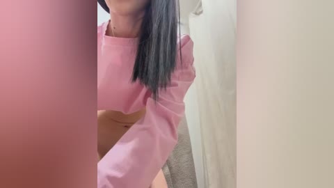 Media: Video of a woman with long black hair, wearing a pink crop top and beige shorts, partially obscured by a person's arm, in a beige room with white curtains.