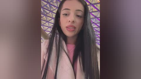 Video of a young, light-skinned woman with long, straight black hair, wearing a pink coat and pink top. She has a neutral expression with slightly parted lips. Background features a purple and white geometric pattern.