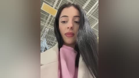 Video of a young woman with long, straight black hair, fair skin, and full lips. She wears a white blazer over a pink top, set against a background of industrial ceiling lights and exposed beams.