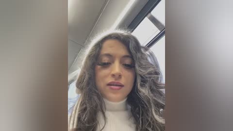 Media: Video of a young woman with long, wavy, light brown hair, wearing a white turtleneck, standing in a modern office with large windows. Her expression is calm, and the background includes office furniture and fluorescent lighting.