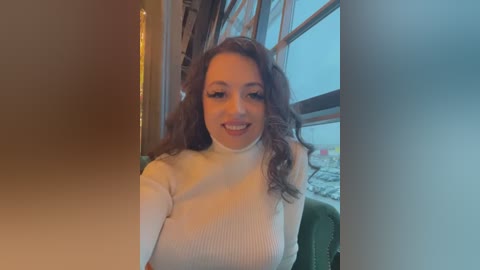 Media: Video of a smiling, light-skinned woman with curly brown hair, wearing a white turtleneck sweater, sitting in a green chair beside a large window with a blurred outdoor view.