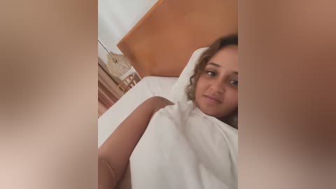 Media: Video of a young woman with curly hair and light brown skin, lying on a bed with a white blanket, looking towards the camera.