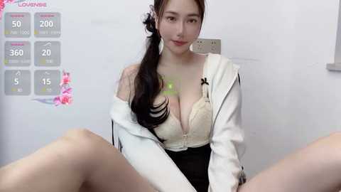 Media: Video of a fair-skinned, Asian woman with long black hair, wearing a white jacket, black skirt, and beige lace bra, sitting with legs spread, in a white room with a calendar and digital clock in the background.