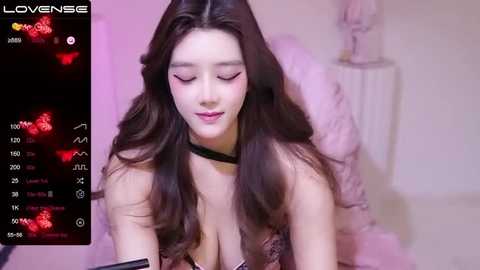 Media: Video of a young Asian woman with long, wavy brown hair, wearing a black choker and a low-cut lace bra, leaning forward, with a pink background.