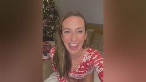 Media: Video of a smiling, light-skinned woman with long brown hair in a red holiday sweater, kneeling on a bed with a decorated Christmas tree and wrapped gifts in the background.