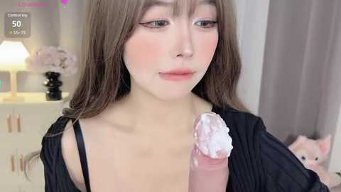 Media: Video of a young Asian woman with fair skin, long brown hair, wearing a black robe, holding a large, erect penis covered in white cream. Background shows a white dresser with a pink teddy bear.