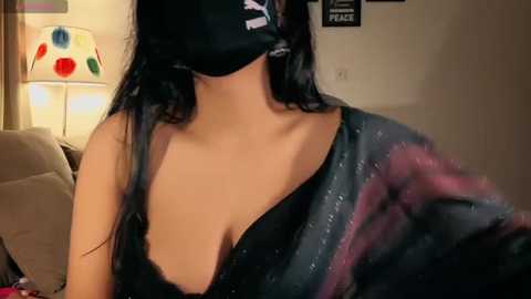 Media: Video of a woman with long black hair, wearing a black mask and black bra, seated indoors. Background features a beige couch, a lamp, and colorful wall art.