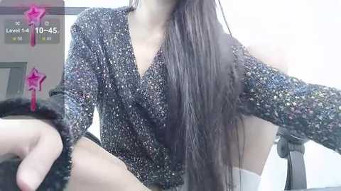 Media: Video of a young woman with long, straight, dark hair, wearing a glittery black long-sleeve top and black fur-trimmed pants, sitting on a chair.