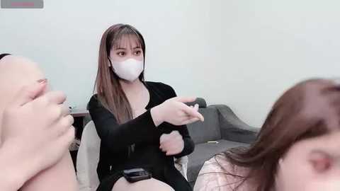 Media: Video of an Asian woman in a black long-sleeve top and white face mask, gesturing with her hands, seated on a gray couch. Two other people, partially visible, are in the foreground.