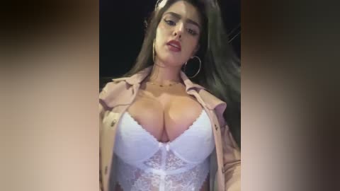 Media: Video of a woman with light brown skin and long, wavy brown hair, wearing a white lace bra, beige blazer, hoop earrings, and a confident expression.