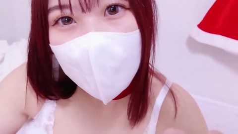 Media: Video of a young woman with long, straight, dark red hair, wearing a white surgical mask, a white spaghetti-strap top, and a red Santa hat. Background is a plain white wall.