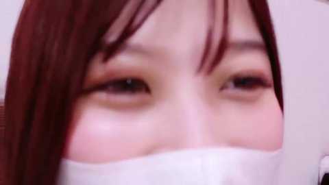 Media: A close-up video of an Asian woman with straight, shoulder-length brown hair, light skin, and light makeup, wearing a white surgical mask. Her eyes are half-closed, and the background is blurred, focusing on her face.