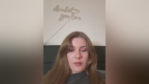 Media: Video of a young Caucasian woman with long, straight brown hair, wearing a dark shirt, looking directly at the camera. Background features a wall with a metallic, cursive \"Believe in yourself\" sign.