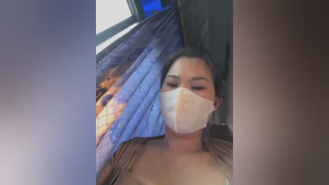 Media: Video of a woman with medium skin tone, dark hair, wearing a white face mask, brown top, lying on a blue and black patterned bedspread.