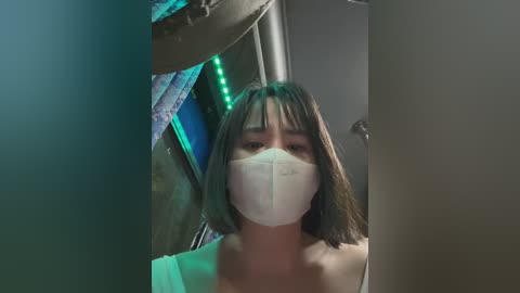 Media: Video of a young Asian woman with shoulder-length black hair, wearing a white face mask, standing indoors next to a blue and green curtain, with a metallic surface in the background.