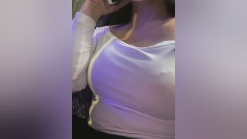 Media: Video of a woman's upper torso, wearing a white, form-fitting top that accentuates her ample cleavage and medium-sized breasts. Her skin tone is light, and she has dark hair partially visible. Background is blurred and dimly lit.