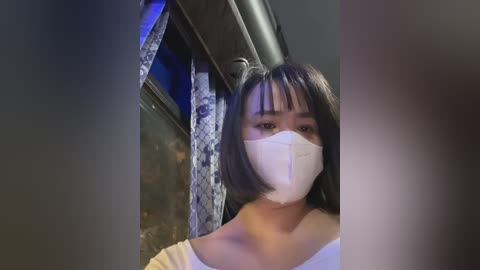 Media: A video of an Asian woman with straight black hair, wearing a white face mask and a light-colored off-shoulder top, standing indoors near a window with blue curtains.