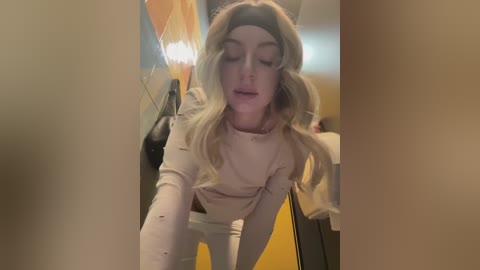 Media: A video of a blonde woman with long, wavy hair, wearing a beige sweater and a headband, smiling and looking down, taken in a dimly lit hallway with a yellow wall and a lamp in the background.