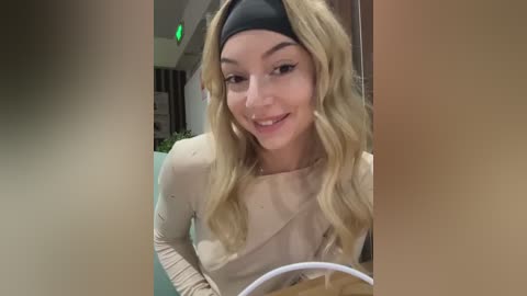 Media: Video of a smiling young woman with wavy blonde hair, wearing a black headband and beige top, seated indoors near a green light and potted plants.