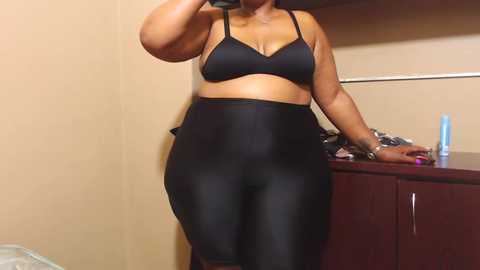 Media: Video of a curvy Black woman with medium skin tone, wearing a black bra and high-waisted shiny black leggings, standing in a dimly lit room with a wooden dresser and a mirror.