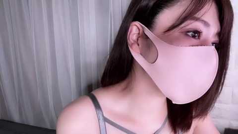 Media: Video of an Asian woman with straight, shoulder-length dark brown hair, wearing a pale pink surgical mask and a grey tank top, standing against a white brick wall with vertical blinds.