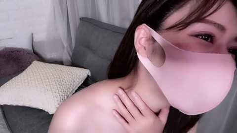 Media: Video of an Asian woman with light skin, wearing a pink face mask and no shirt, lying on a gray couch, with a textured cream pillow and sheer curtains in the background.