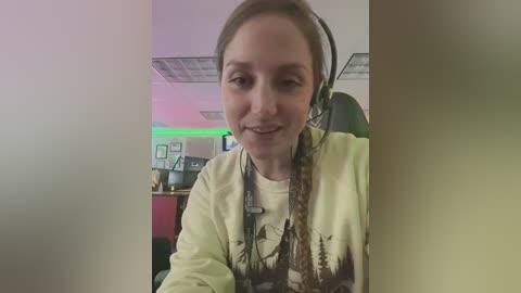 Media: A video of a young woman with light skin, brown hair, and a slight smile, wearing a headset, green neon lights, and a t-shirt with a nature print, sitting in a modern office with framed pictures on the wall.