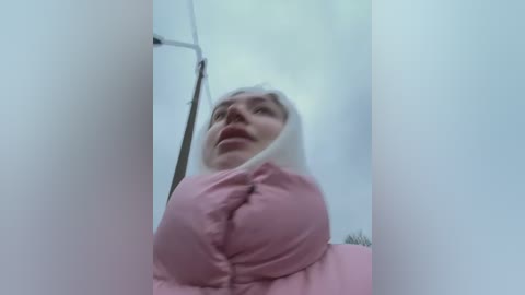 Media: A low-angle, blurry video of a woman with fair skin, wearing a pink puffy jacket and a white hood, standing in a white-tiled bathroom.