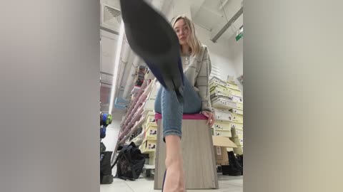 Media: Video of a blonde woman in a grey cardigan and jeans, standing on a wooden stool in a cluttered office or storage room.