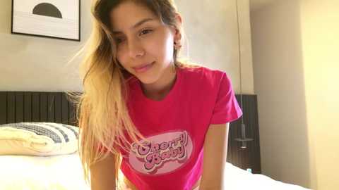 Media: Video of a young woman with long blonde hair, wearing a pink \"Dairy Queen\" t-shirt, sitting on a bed in a brightly lit, minimalist bedroom.