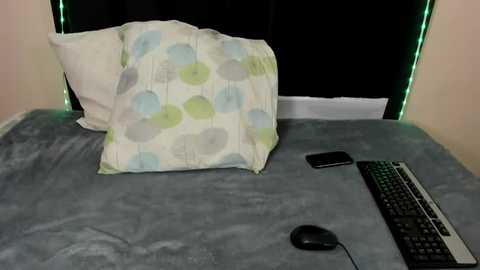 Media: Video of a cozy bedroom with a white pillow adorned with light blue and green circles, resting on a grey bedspread. A black computer mouse and keyboard are positioned on the bed, with a smartphone lying nearby. Green LED strip lights frame the bed.