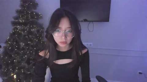 Media: A video of a young Asian woman with long black hair and glasses, wearing a black top with cut-outs, sitting in front of a lit Christmas tree and flat-screen TV.