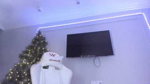 Media: Video of a modern living room with a Christmas tree adorned with white lights and a white Santa hat on a white pillow, a flat-screen TV mounted on a gray wall, and blue LED strip lights along the ceiling.