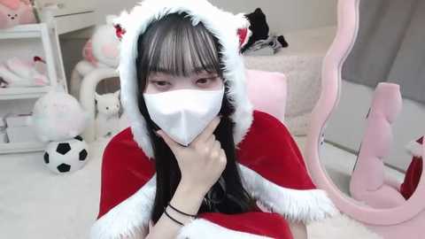 Media: A young Asian woman with long black hair, wearing a white face mask, red Santa hat, and red cape, sits in a room filled with plush toys and a pink gaming chair.