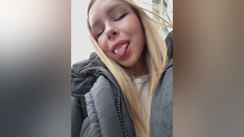 Video of a young blonde woman with long hair, wearing a dark puffer jacket, sticking out her tongue while looking down, taken from an out-of-focus car window.