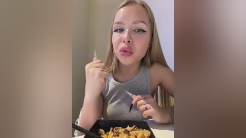 A video of a young blonde woman with fair skin, wearing a gray sleeveless top, eating cereal from a black bowl with a black spoon. She has a playful expression, with her lips puckered and eyes closed.