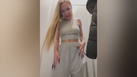 Media: Video of a slim, blonde young woman in a beige crop top and baggy pants, standing in a dimly lit hallway, partially obscured by shadows.