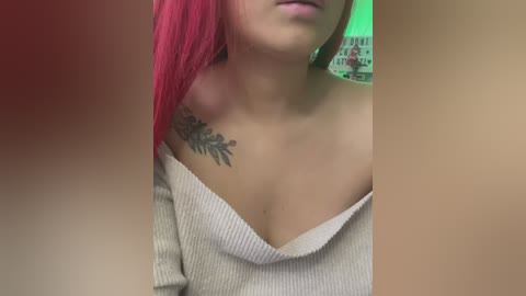 Media: Video of a woman with bright pink hair, wearing a beige off-shoulder ribbed sweater, showing a detailed tattoo on her collarbone. Background features green wall art and blurred objects.