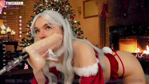 Media: A video of a woman with long silver hair, wearing a revealing Santa costume, licking a large, translucent dildo, in a cozy, lit living room with a decorated Christmas tree and fireplace.