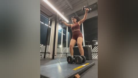 Media: Video of a determined woman with dark skin and curly hair, lifting weights in a modern gym with industrial decor. She's wearing a burgundy sports bra and shorts.