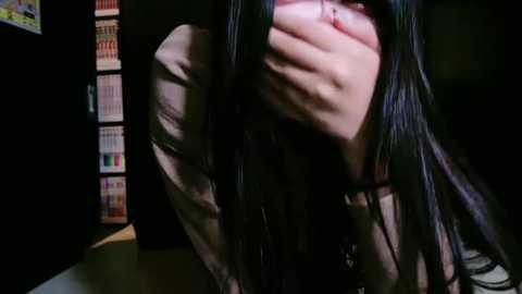 Media: Video of a woman with long black hair, covering her mouth, sitting at a desk. Background features a dark room with a bookshelf and colorful file folders.