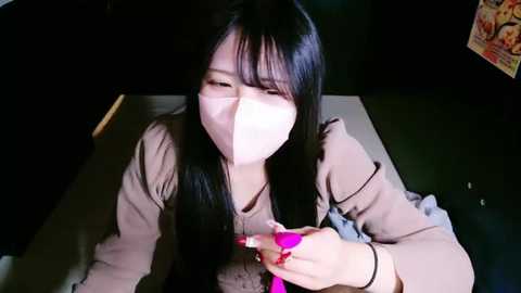 Media: Video of an Asian woman with long black hair and fair skin, wearing a beige top, face mask, and pink manicured nails, holding a pink smartphone in dimly lit indoor setting.