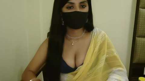 Media: Video of a South Asian woman with long black hair, wearing a black mask, yellow sari, and a revealing blue bra, seated indoors with a beige wall background.