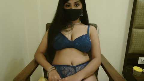 Media: A video of a South Asian woman with long black hair, wearing a blue lace bra and panties, seated in a wooden chair against a beige wall.